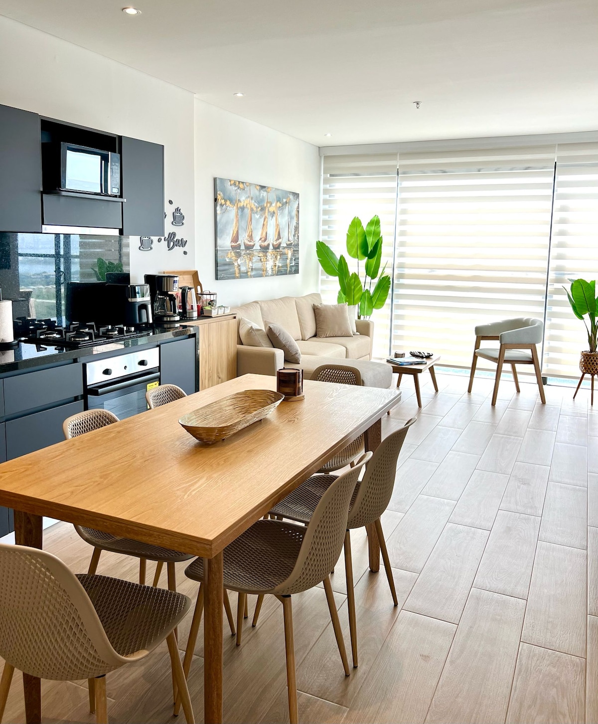 A modern kitchen area is presented with an open layout, featuring a wooden dining table and six grey chairs. A cozy living space is visible, equipped with a comfortable sofa and decorative plants. Light streams through large windows, complemented by soft, airy window treatments.
