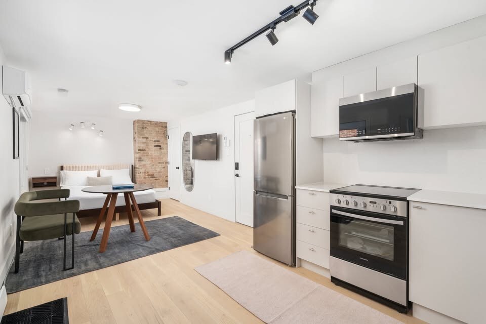 A modern open-concept living space features a well-equipped kitchen with stainless steel appliances. A wooden dining table is complemented by two green chairs, while a cozy bed can be seen in the background, creating a homely environment.