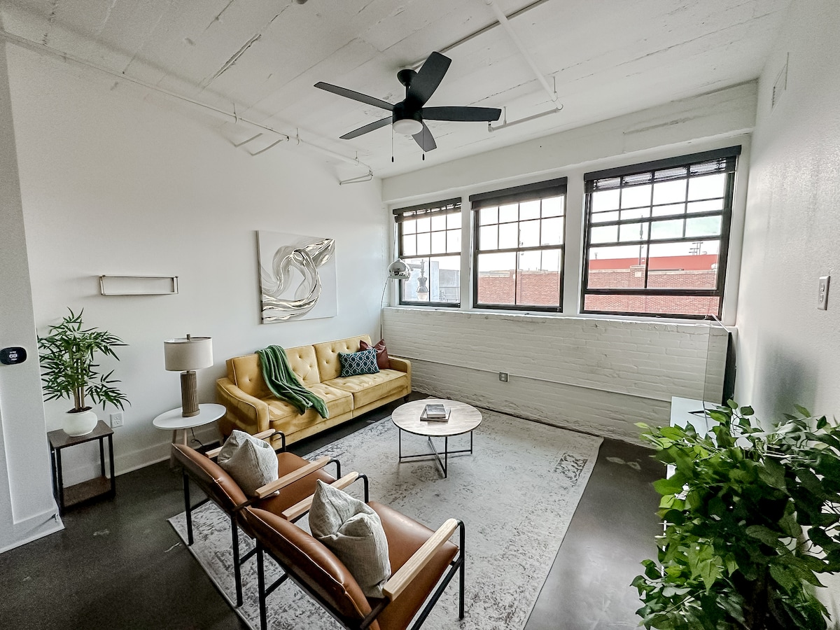 A bright and airy living room is showcased, featuring large windows that allow natural light to flow in. A comfortable yellow sofa is paired with two chairs and a round coffee table. Decorative plants and artwork add a touch of style to the space.