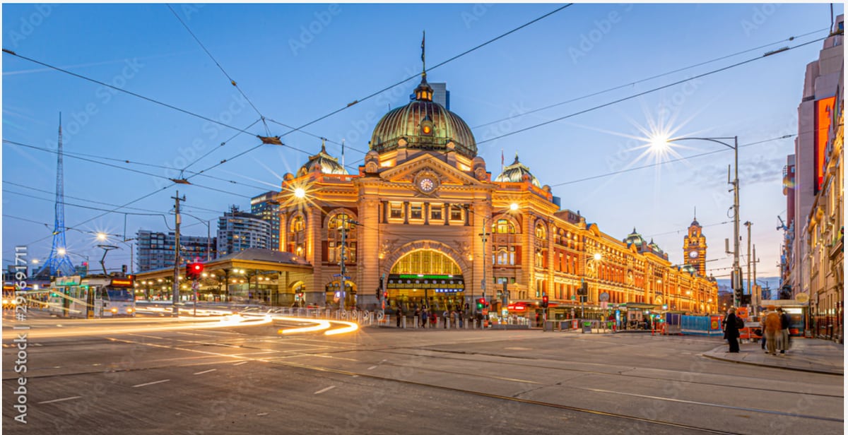 The iconic Flinders Street Station is illuminated as dusk falls, showcasing its grand architectural features. The building's distinct dome and arched entrance are highlighted by warm lights, while trams and vehicles navigate the bustling streets in the foreground.