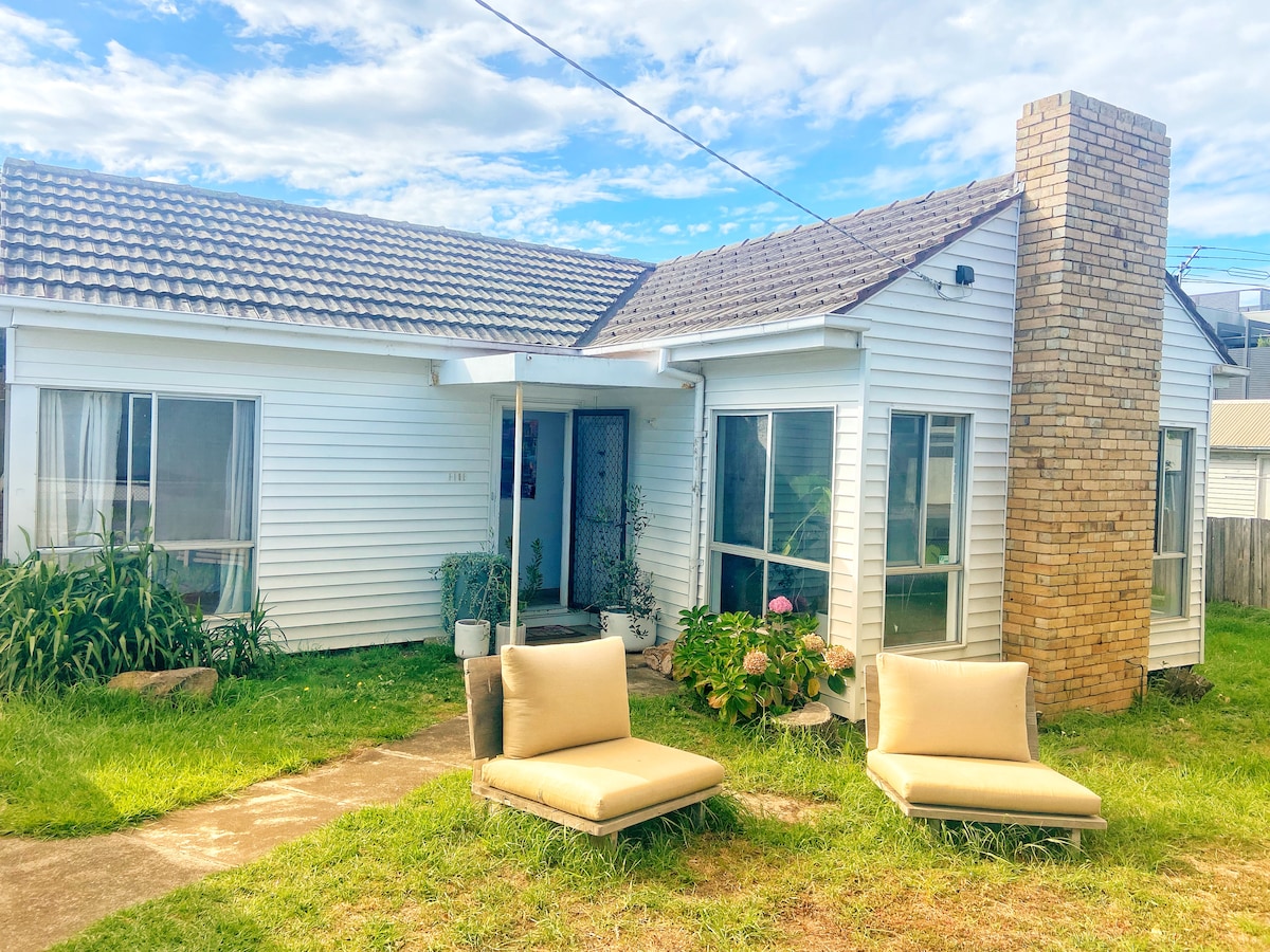 The charming exterior of the cottage is showcased, featuring light-colored siding and a prominent brick chimney. Two cushioned seats are placed on a grassy area, inviting relaxation. Abundant greenery surrounds the entrance, enhancing the cottage's peaceful ambiance.