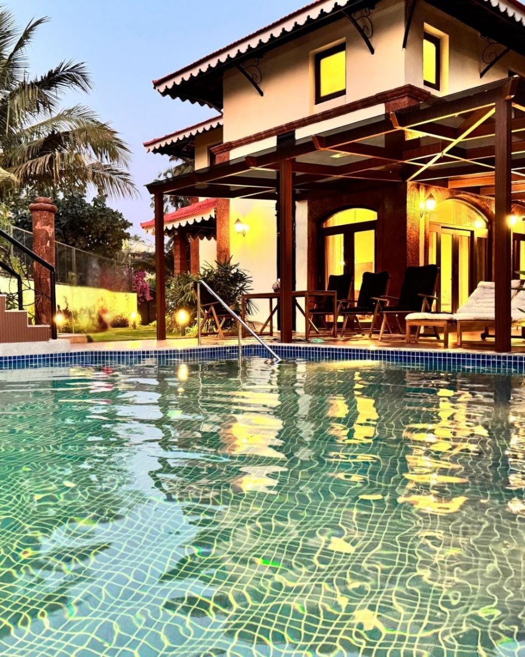 The image captures a private pool reflecting warm lights from the villa’s exterior. The villa, featuring a blend of traditional and modern architecture, is framed by palm trees. Outdoor seating is arranged on a deck, providing a welcoming space for relaxation beside the water.