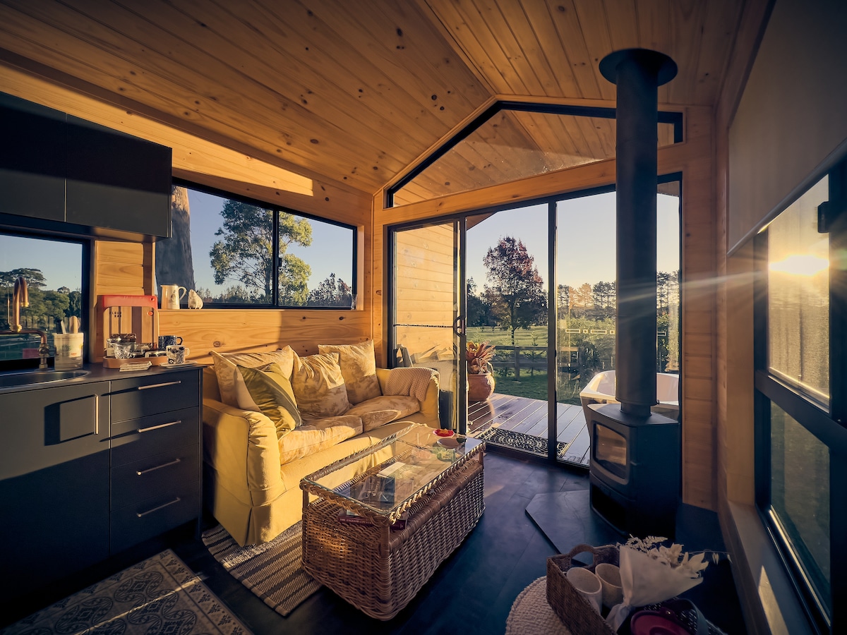 A cozy living area is highlighted by warm wood paneling and large windows that provide views of the surrounding farmland. A comfortable sofa is paired with a glass coffee table, while a sleek wood stove stands in one corner, adding a touch of warmth to the space.