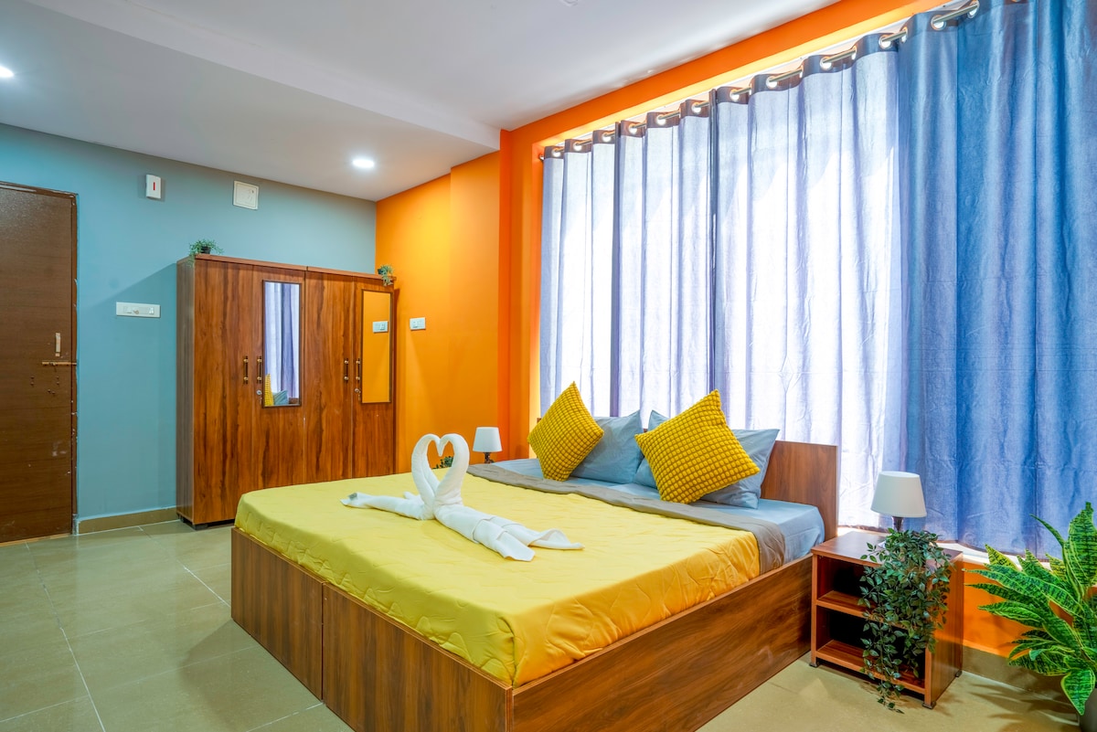 The studio room features a spacious bed adorned with yellow bedding, complemented by two decorative pillows. Large windows are draped with light blue curtains, allowing natural light to brighten the space. A potted plant adds a touch of greenery, enhancing the overall inviting ambiance.