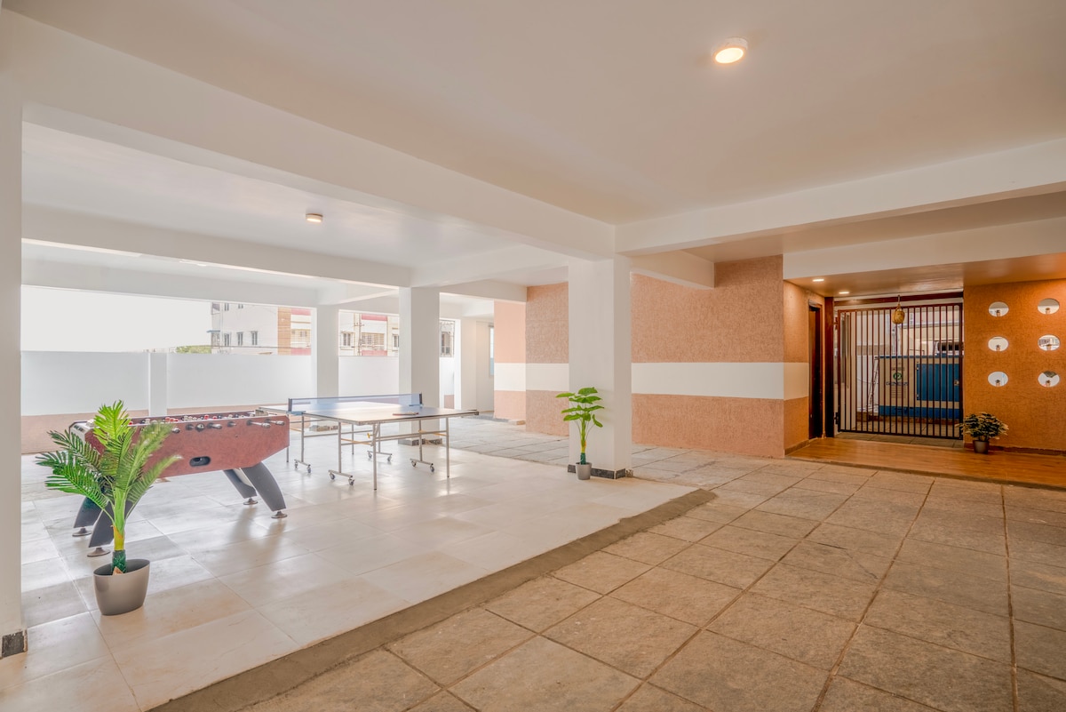 An open recreational area features a table tennis setup beside a foosball table, both set on tiled flooring. Potted greenery adds a refreshing touch, while bright lighting enhances the spacious atmosphere. The entrance in the background leads to additional shared spaces.