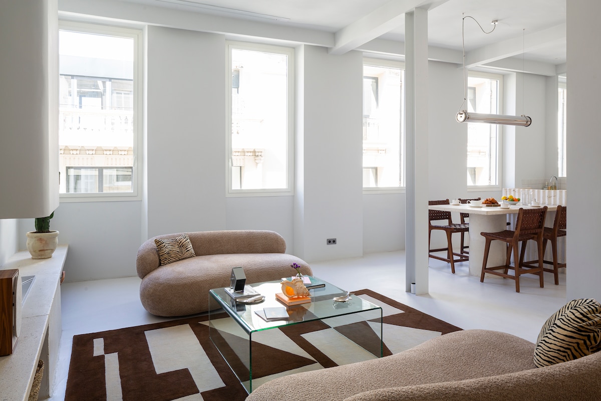 A bright living space is showcased with large windows allowing natural light to fill the room. Modern, curved sofas face a glass coffee table, which holds various decor items. A dining table with wooden chairs can be seen in the background, highlighting an open layout.