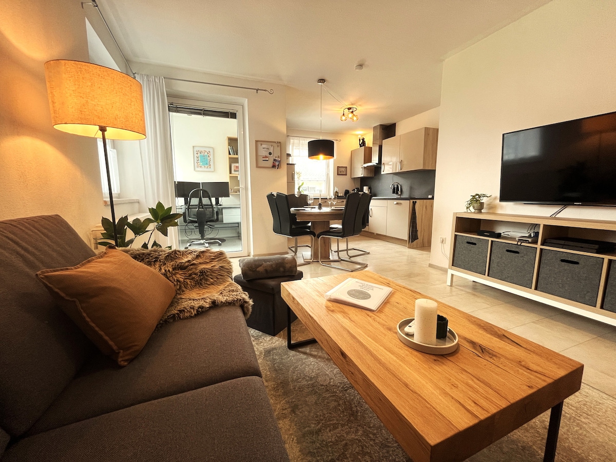 A cozy living area features a comfortable sofa adorned with decorative pillows, alongside a wooden coffee table with a candle and a book. A dining set is visible in the background, accompanied by a modern kitchen area and large windows allowing natural light.