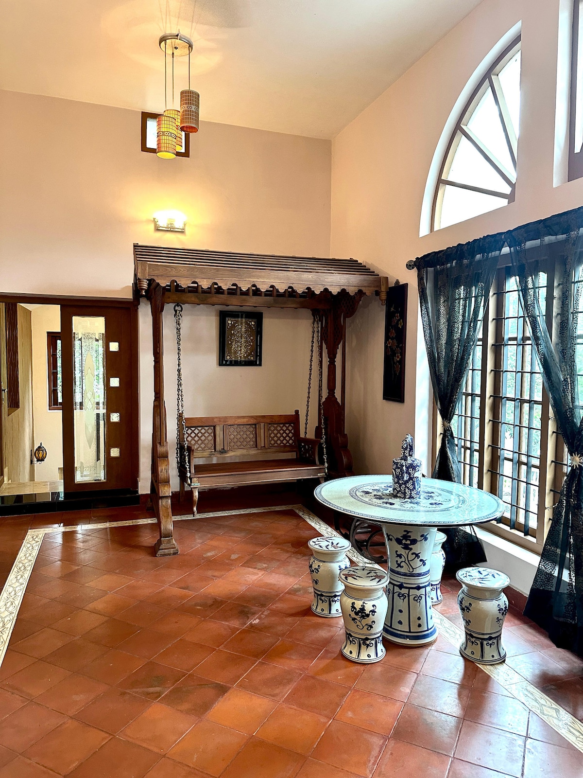 A welcoming entryway features a traditional wooden swing, complemented by a round table and matching stools with intricate designs. Large arched windows allow natural light to fill the space, while dark curtains frame the windows and add a touch of elegance.