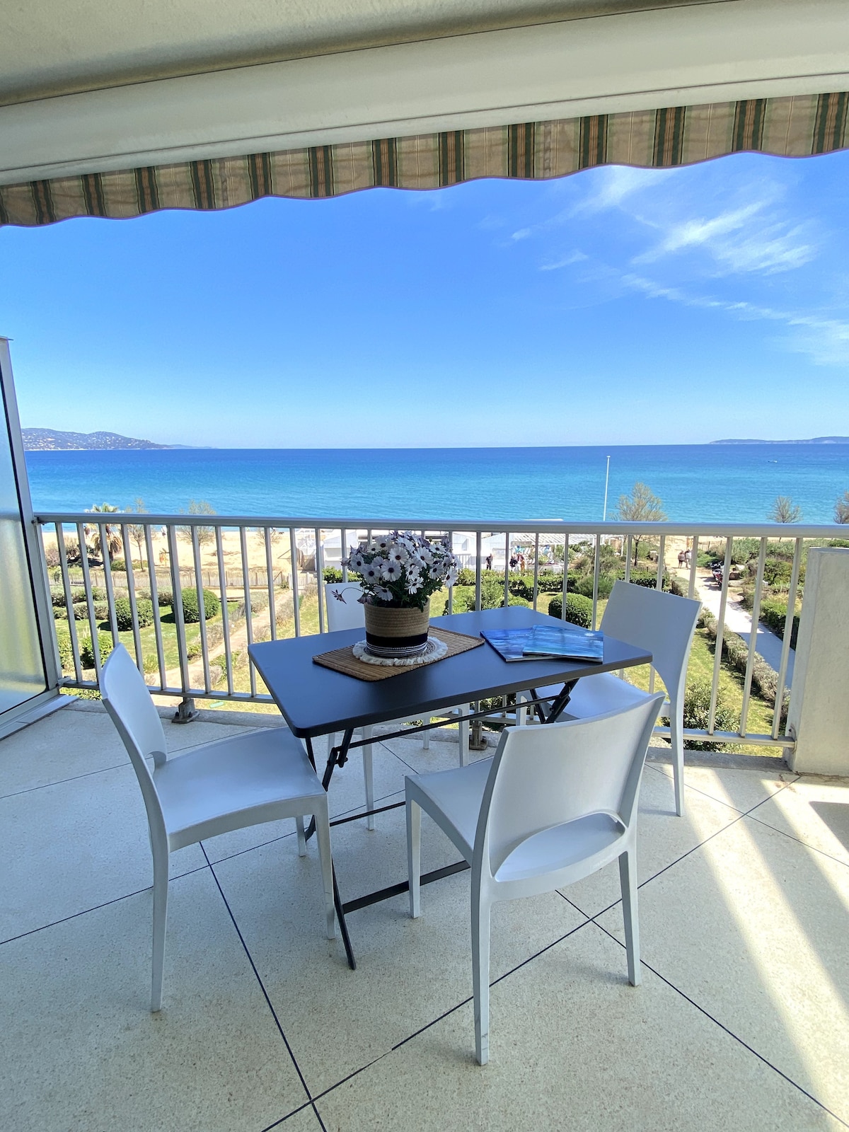 A balcony table set for four is presented with a vase of flowers, overlooking a serene view of the sea. The bright blue waters stretch into the horizon, with a clear sky enhancing the scene. A sunshade provides comfort and shade for outdoor enjoyment.