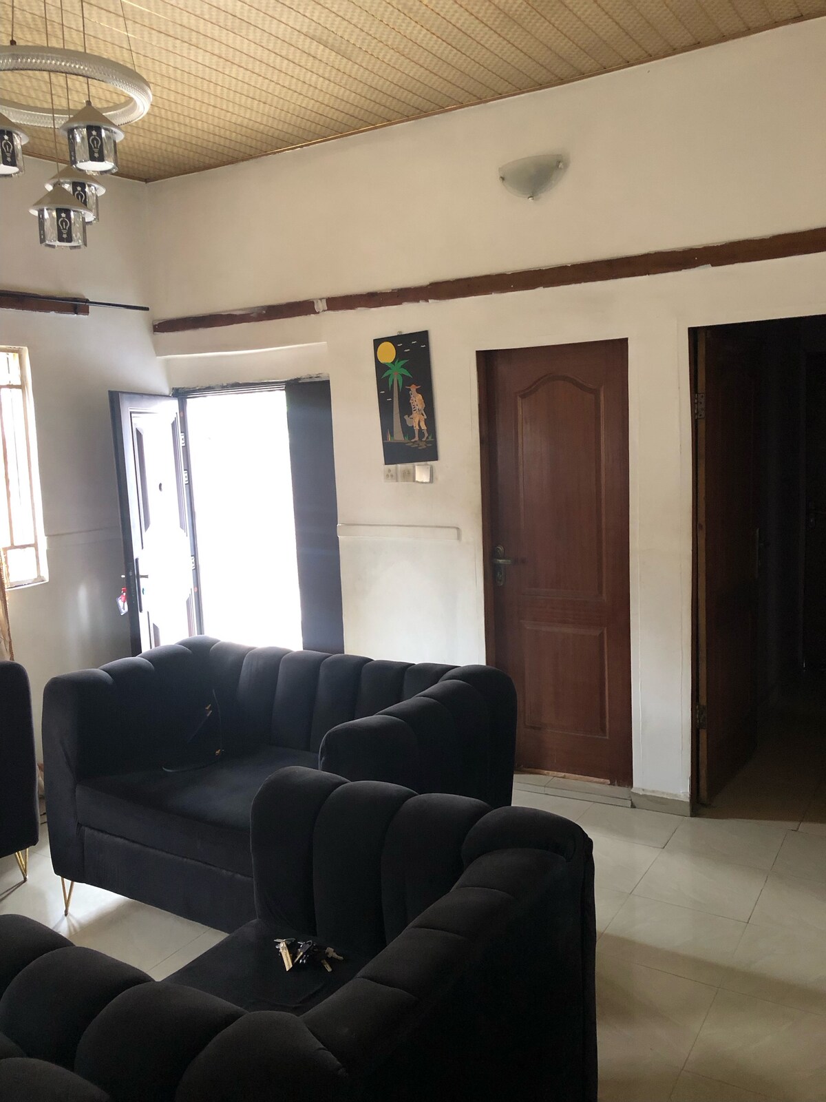 A spacious living area features stylish black sofas arranged invitingly. Natural light filters in through large windows, illuminating the room. A simple artwork is displayed on the wall, contributing to the overall design. The entrance to the room is accessed through a door on the left.
