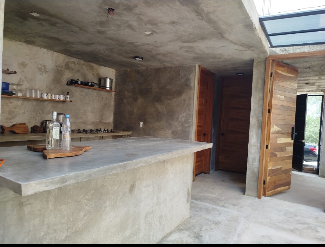 An open kitchen area features a concrete countertop with wooden shelving displaying glassware. A gas stove is positioned on the back wall, while two wooden doors lead to adjacent spaces. Natural light filters in through a skylight, enhancing the rustic design.
