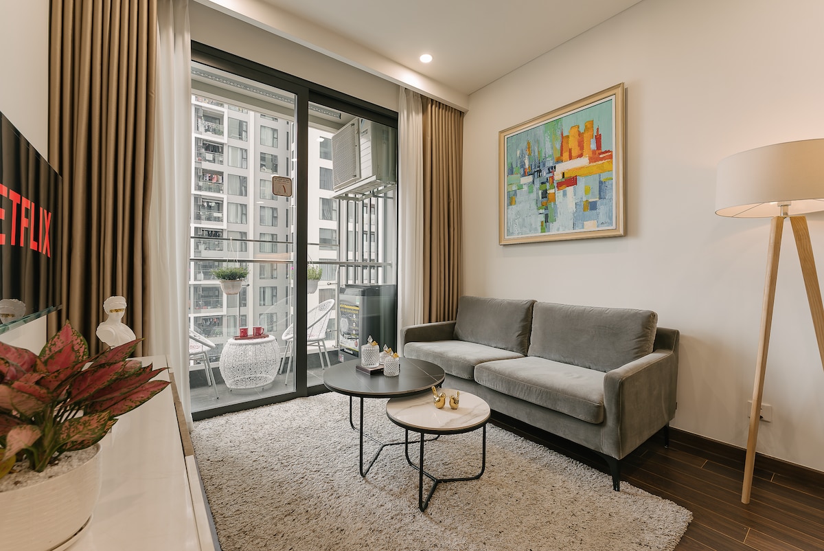 A comfortable living area is presented with a gray sofa positioned in front of a glass door leading to a balcony. Two small tables are arranged on a soft rug, while a colorful artwork adorns the wall above. Natural light filters in through the large windows.