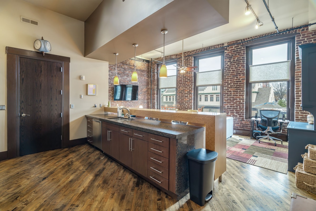 The kitchen area features modern wooden cabinetry and countertops, illuminated by pendant lighting. Large windows offer natural light, highlighting the brick wall and wooden flooring. A seating area is visible, with a variety of textures in the carpet, while a workspace is set near the windows.