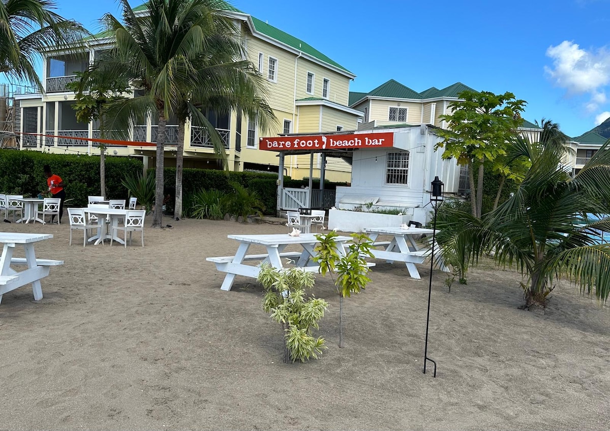 A beachfront bar, known as Barefoot Beach Bar, is featured under a red sign. Multiple white picnic tables are arranged on the sandy beach, surrounded by palm trees. A light green shrub adds a vibrant touch to the sandy area near the bar.