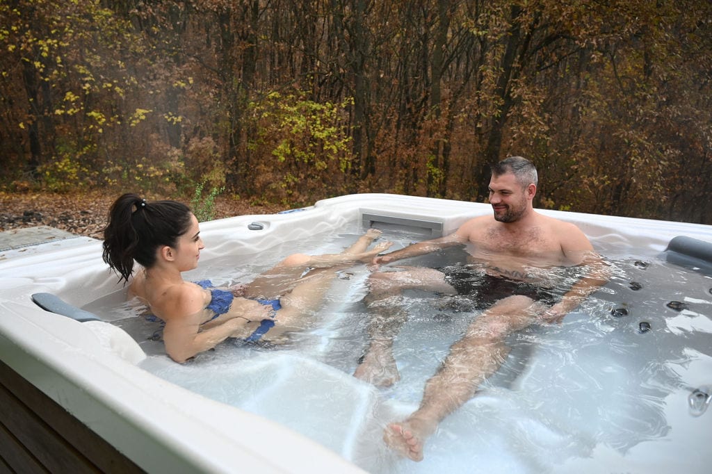 A relaxing hot tub is surrounded by a wooded area showcasing autumn foliage. Bubbles rise around two individuals enjoying the warm water, which is set against a tranquil natural backdrop. Soft lighting suggests a calm atmosphere for unwinding.