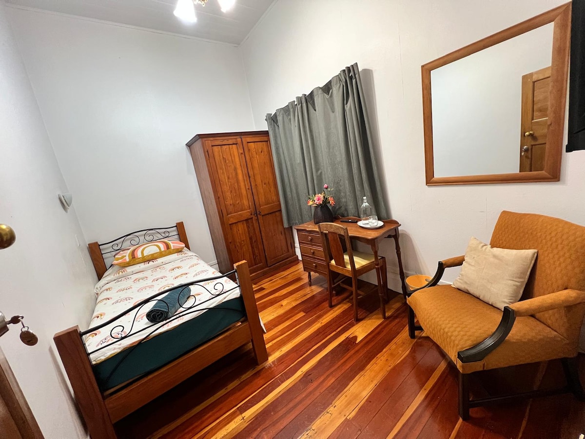 An intimate single room features a cozy bed with decorative bedding. A wooden wardrobe and a simple desk with a chair are present. The room is illuminated by natural light filtering through gray curtains, while warm wooden floors add to the inviting atmosphere.