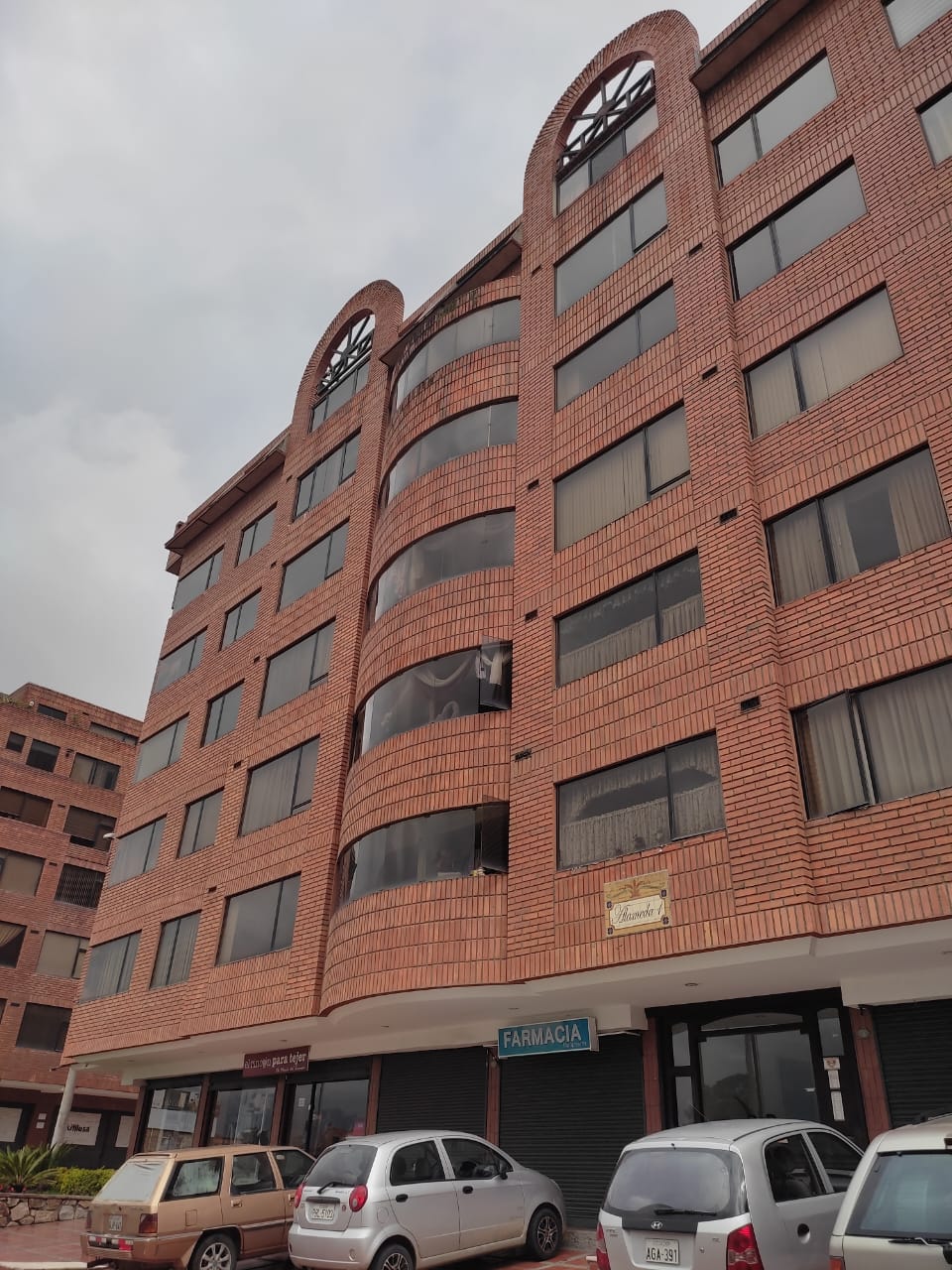 A multi-story brick building is showcased with large, curved windows. Ground-level commercial spaces are visible, including a pharmacy sign. A few parked cars line the street in front, contributing to the urban environment. The sky is overcast, adding a mellow ambiance.