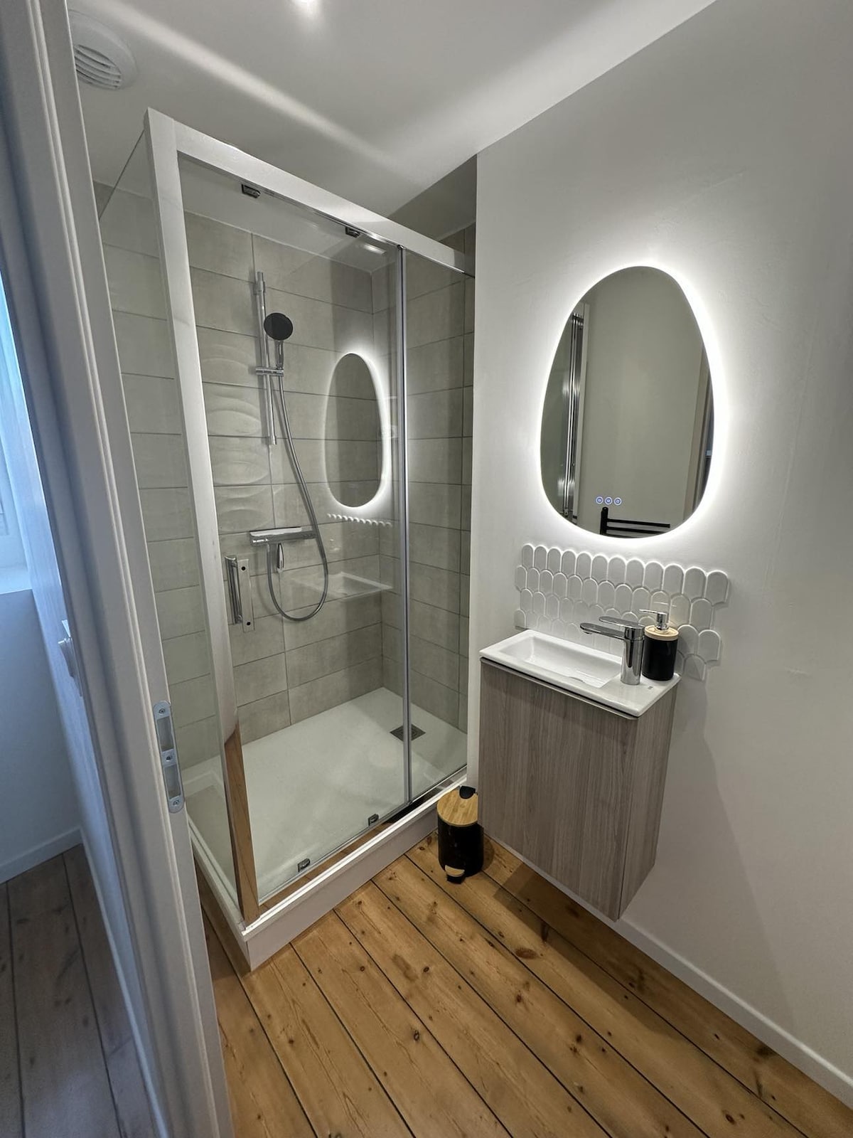 A modern bathroom is visible, featuring a spacious walk-in shower enclosed by glass. A large oval mirror reflects soft lighting above a compact sink, which is placed against a light wood cabinet. Natural light enters through a small window, enhancing the room's fresh aesthetic.
