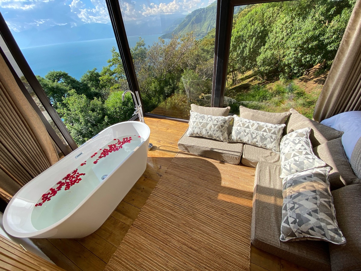 A modern living space features a comfortable sofa set with patterned cushions, situated next to a freestanding bathtub filled with rose petals. Large glass windows provide sweeping views of the natural surroundings and the lake, inviting ample natural light into the room.