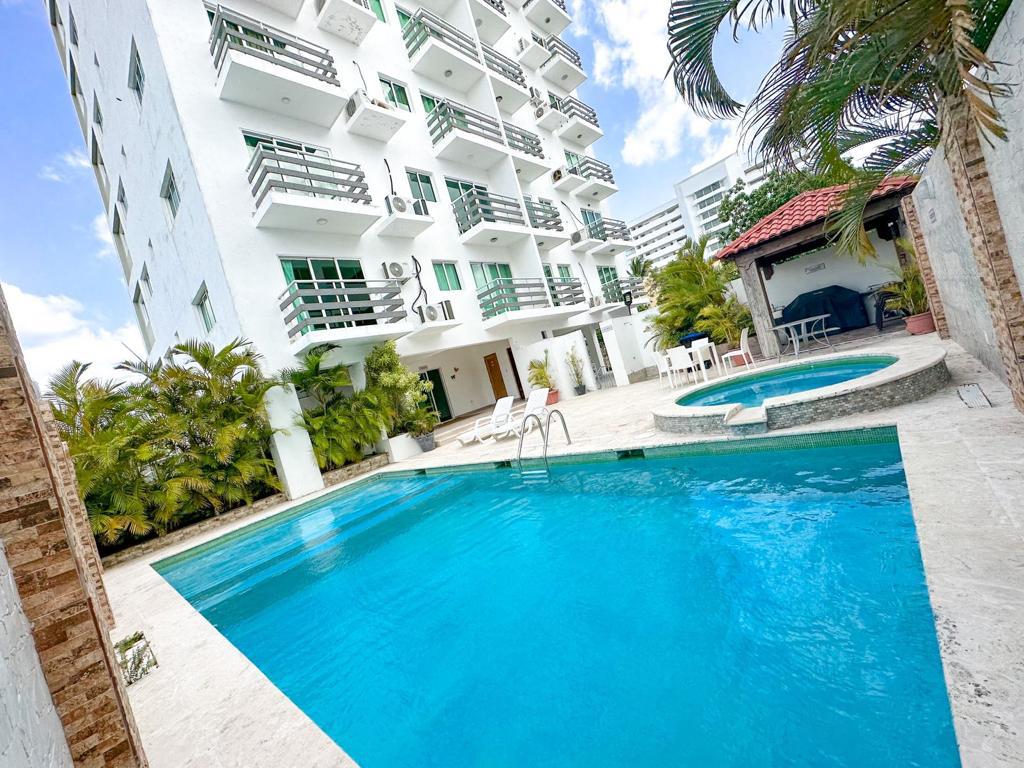 An inviting outdoor area features a large swimming pool with clear blue water, surrounded by lush greenery. A smaller hot tub is situated nearby. The building's spacious balconies overlook the pool, while a shaded seating area offers relaxation opportunities.