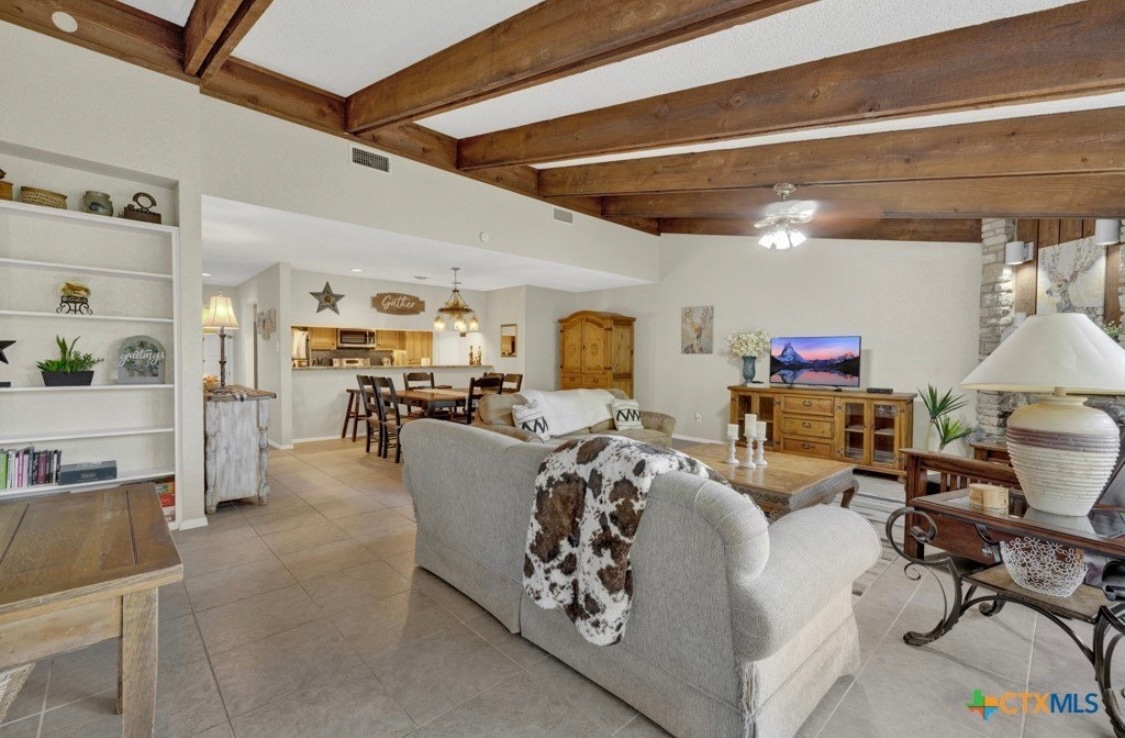 An open concept living area features a spacious sectional sofa adorned with decorative pillows. Exposed wooden beams span the ceiling, and a large TV is positioned on a wooden console. A dining area is visible in the background, accompanied by additional seating options.