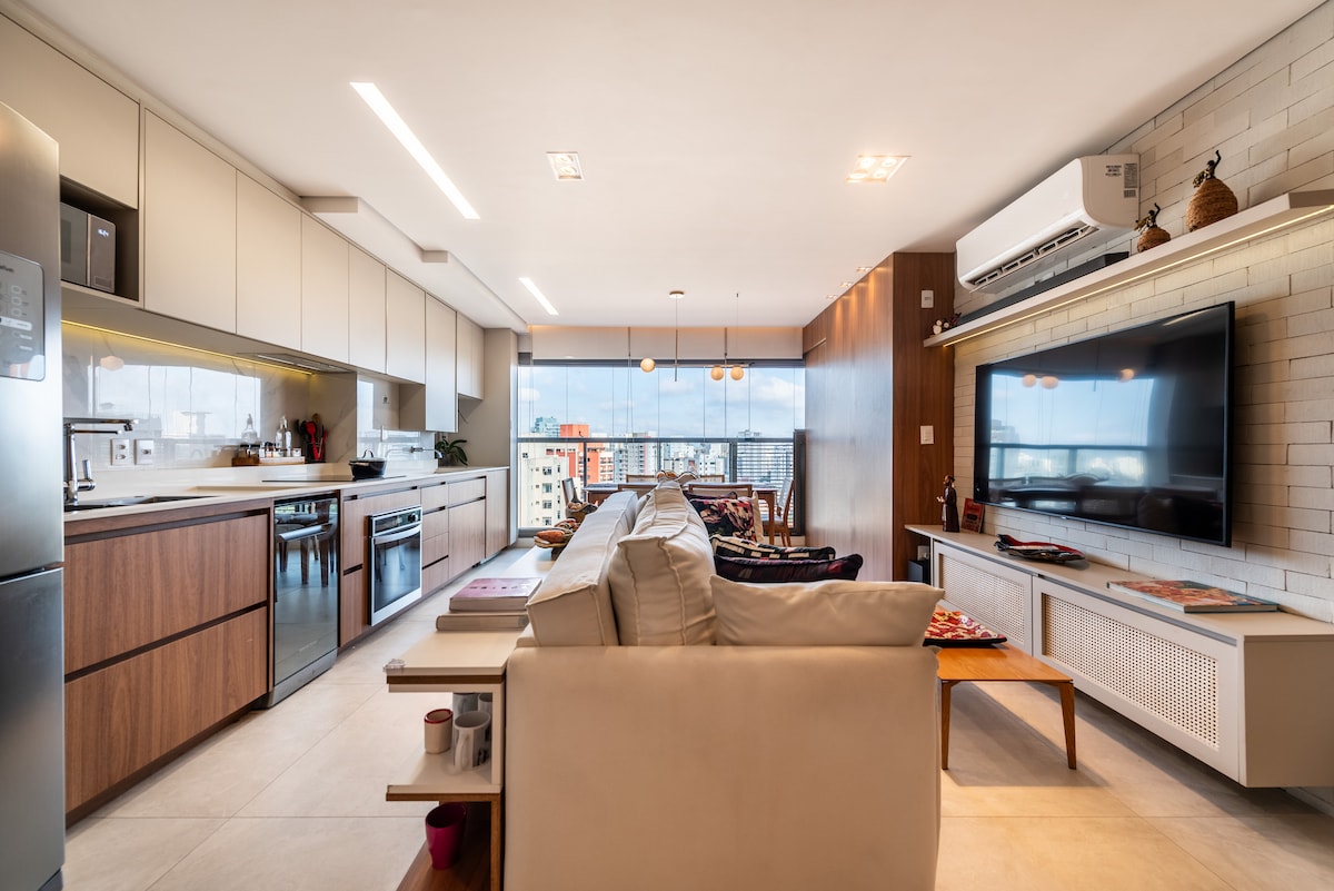 An open-plan living area reveals a modern kitchen with sleek cabinetry and stainless steel appliances. A comfortable seating arrangement faces a wall-mounted television. Large windows provide a view of the city, allowing ample natural light to fill the space, highlighted by minimalist decor.