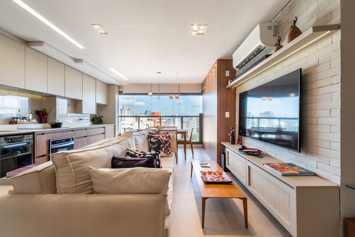 An open living area is presented, featuring a comfortable sectional sofa facing a wall-mounted television. A wooden dining table is seen in the background, surrounded by four chairs. Natural light fills the space through large windows, while modern cabinetry lines the kitchen area.
