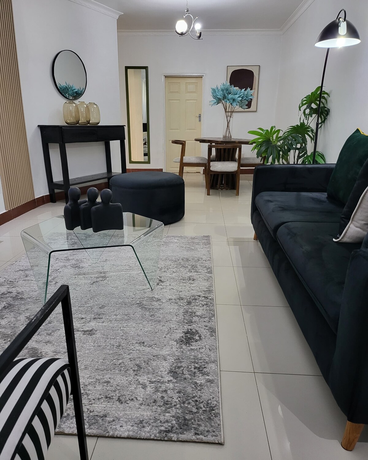 A modern living area features a dark green sofa and a glass coffee table atop a textured rug. Decorative items are displayed on a black console table, while a small dining area with wooden chairs is visible in the background, complemented by potted plants.