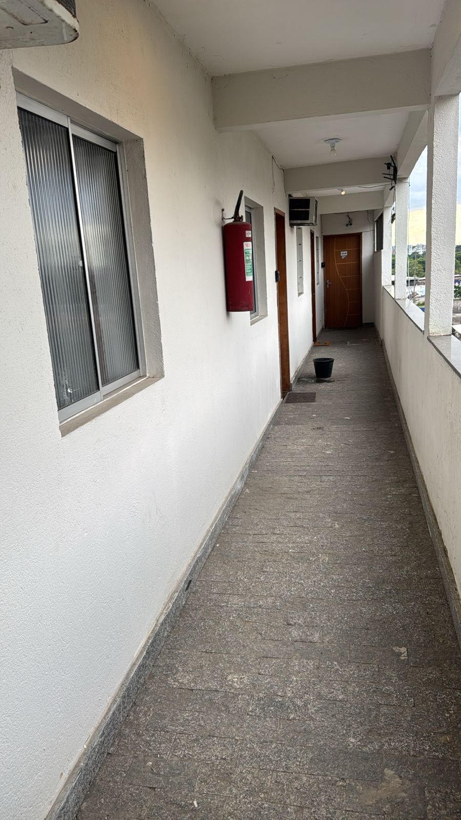A narrow hallway is depicted, featuring a light-colored wall and a concrete floor. A window is located on the left side, allowing natural light to fill the space. Two doors are visible along the right side, with a fire extinguisher mounted near one door. A small black bin is placed on the ground.