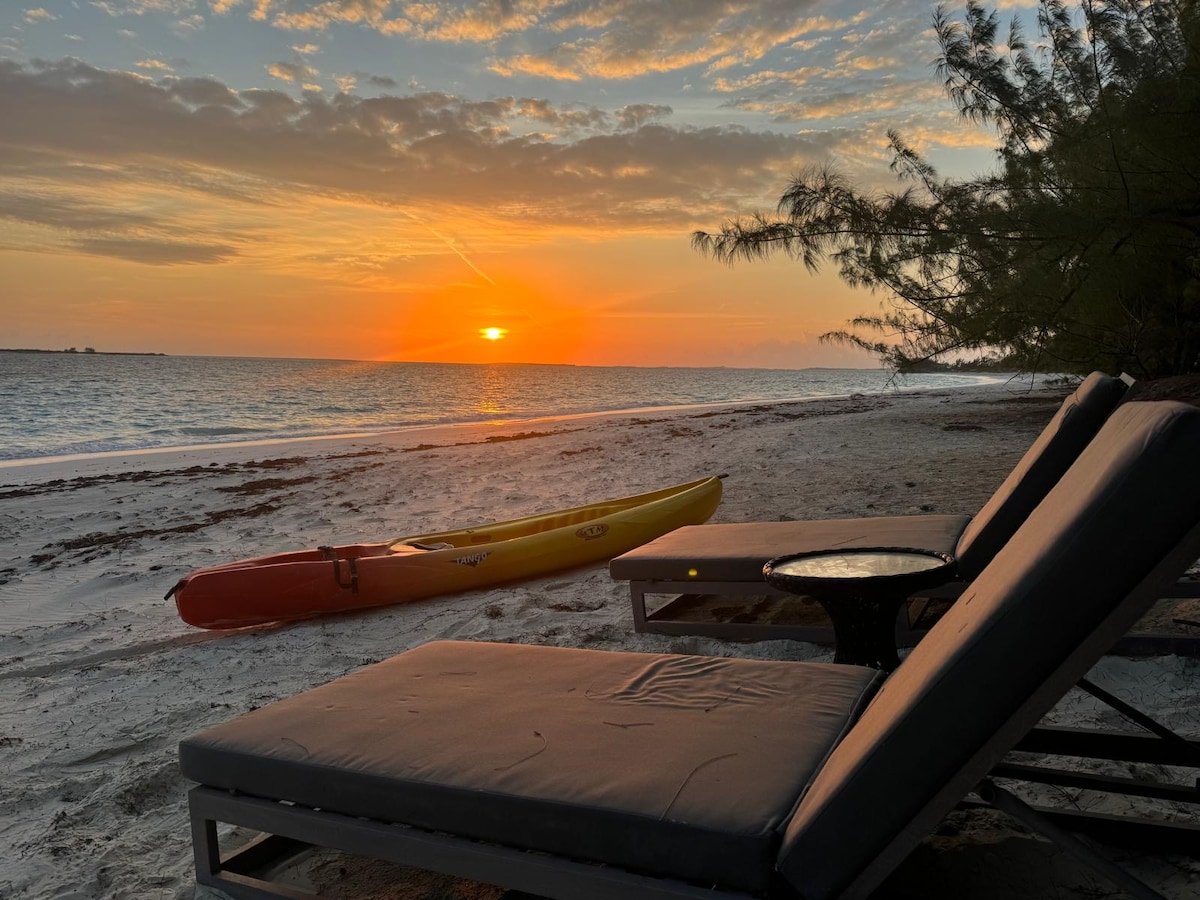 Lounge chairs are positioned on a sandy beach, overlooking calm waters at sunset. A kayak rests on the shore, and the sky is painted in soft hues of orange and pink, reflecting a serene atmosphere around the coastal retreat.