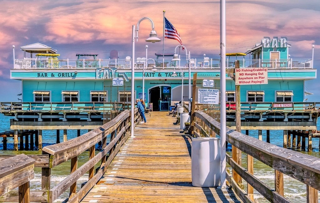 A wooden pier extends over calm water, leading to a vibrant blue building labeled 'Bar & Grill.' The structure features outdoor seating and an American flag flying above. Soft clouds are visible in the sky, creating a colorful backdrop.