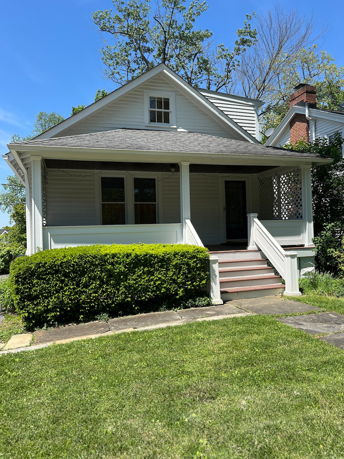A light-colored cottage with a covered front porch and stairs leading to the entrance. Lush greenery surrounds the home, and a manicured lawn is visible in the foreground. A brick chimney can be seen peeking from the roof, indicating classic architectural details.