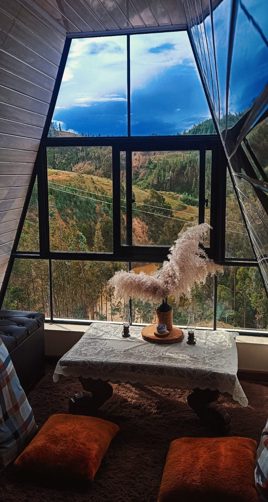 A cozy corner features a low table adorned with decorative items, surrounded by plush cushions. Large windows offer expansive views of rolling hills and lush greenery, allowing natural light to illuminate the space and create a serene connection with the outdoors.