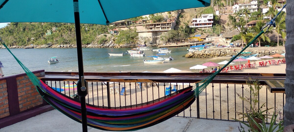 A colorful hammock is suspended between two posts, overlooking a serene beach scene. The water is calm with several small boats anchored nearby. Beach umbrellas and lounging chairs dot the sandy shore, framed by lush hillside and charming beachfront buildings.