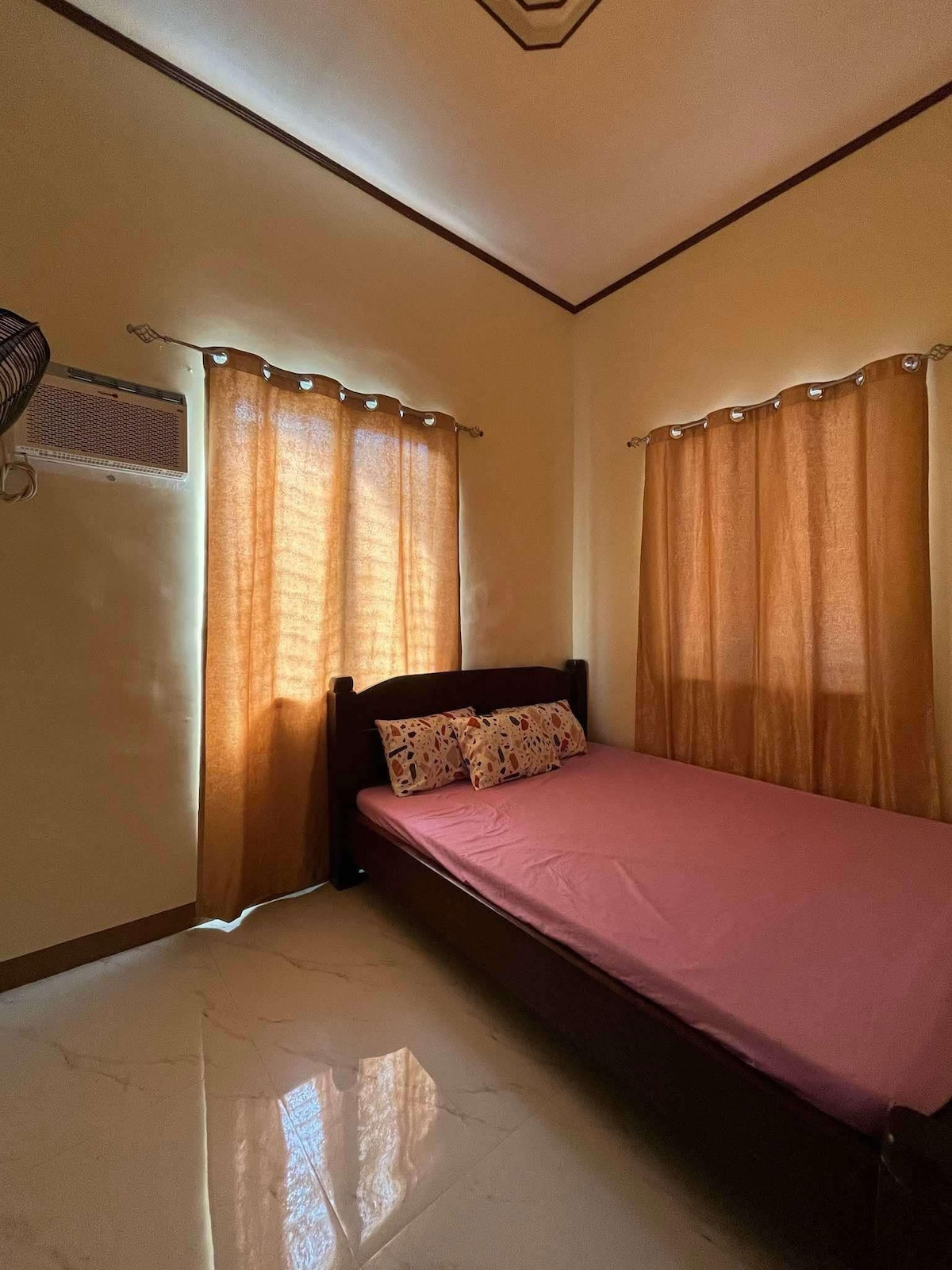 A neatly arranged sleeping area features a spacious bed with a light pink mattress cover. Natural light filters through two flowy brown curtains over the windows, enhancing the warm ambiance. An air conditioning unit is mounted on the wall, providing comfort during warmer months.