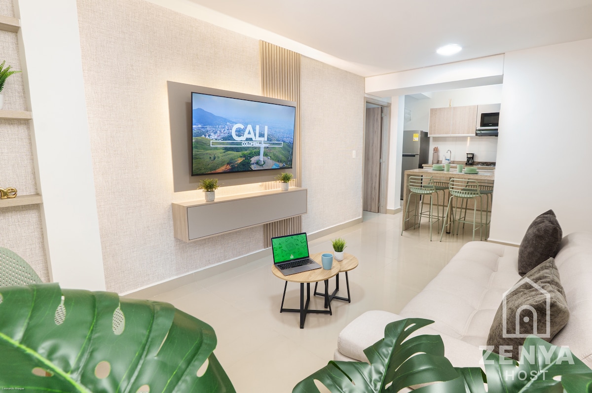 A modern living area features a large television mounted on a light-colored wall. A small coffee table with greenery sits in front of a comfortable seating arrangement. In the background, a kitchenette is visible, with a dining table and chairs contrasting against the neutral tones.