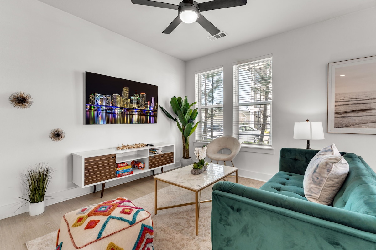 A modern living area is presented with a plush green sofa and a decorative coffee table. The wall-mounted television displays a vibrant city skyline at night. Large windows allow natural light to brighten the space, complemented by decorative plants and stylish artwork.