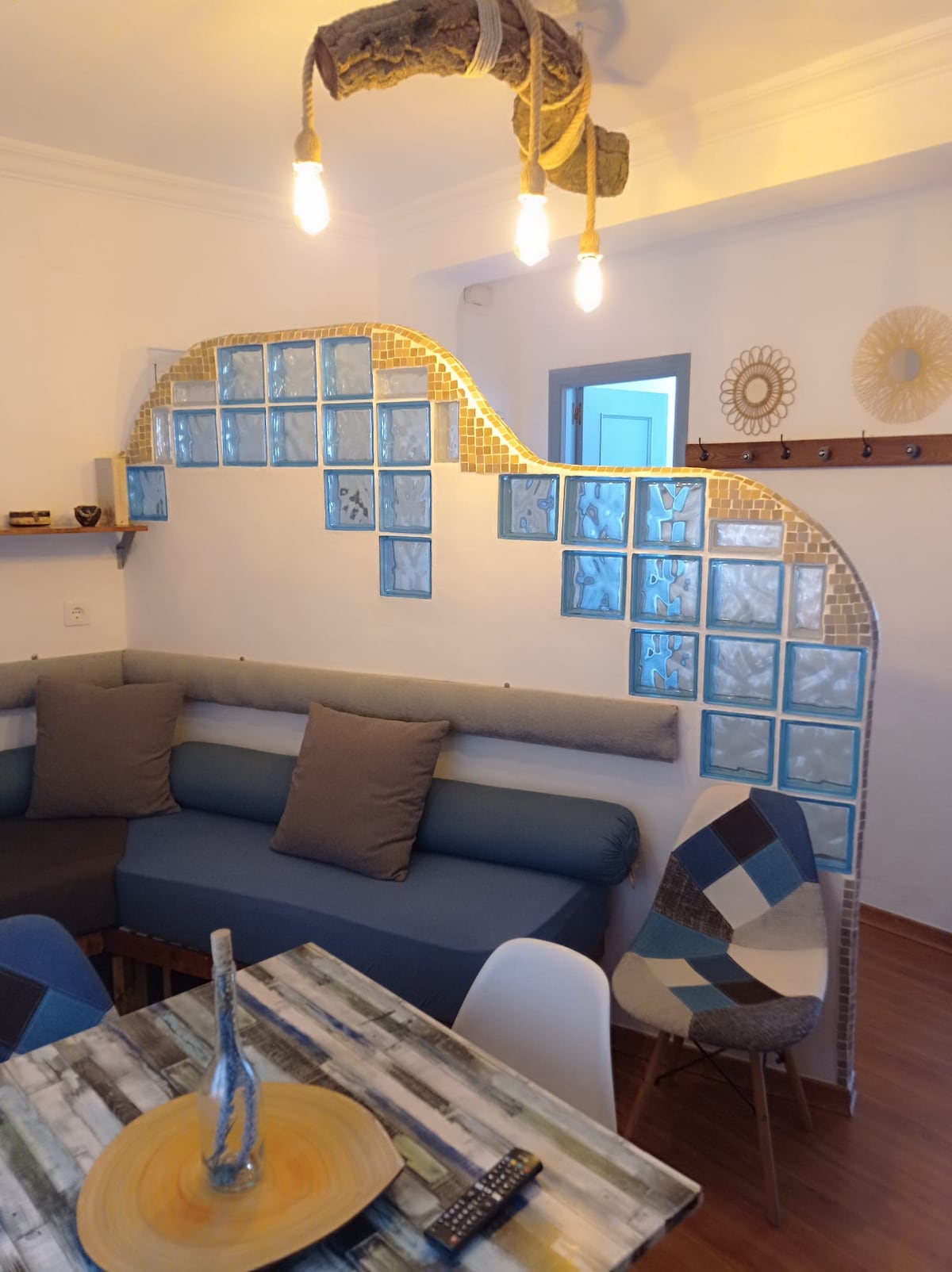 A comfortable seating area is highlighted with a curved wall featuring blue glass tiles. A wooden table is paired with modern chairs, and warm lighting is provided by hanging bulbs above. A textured wall and decorative items add character to the space.