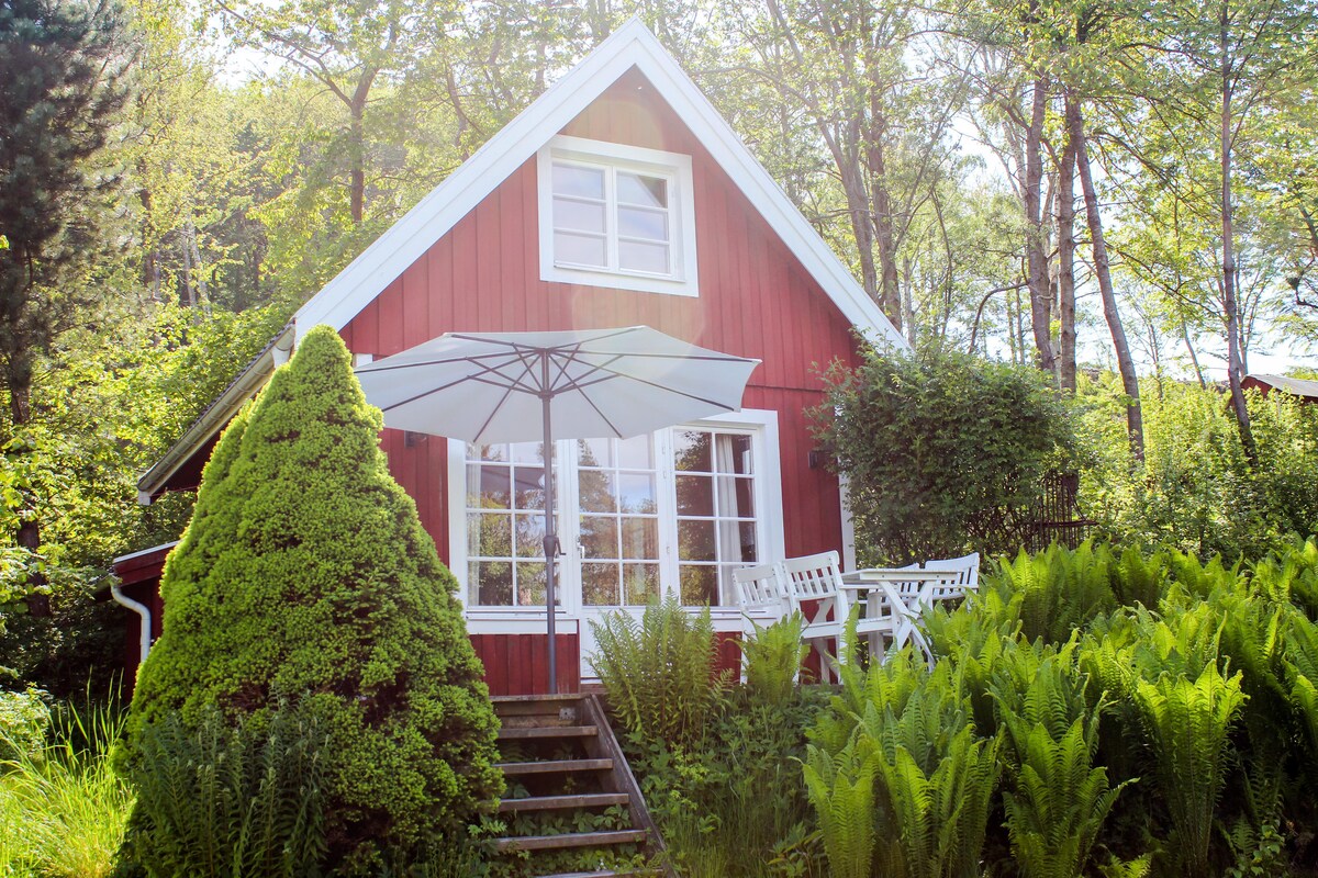This charming red cottage is enveloped by lush greenery and trees, creating a serene atmosphere. A spacious umbrella shades a small outdoor seating area, featuring white chairs and a table, inviting relaxation amidst nature.
