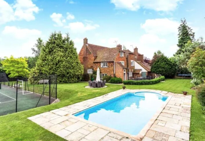 The exterior view displays the historic farmhouse surrounded by lush greenery and mature trees. A heated pool is situated in the foreground, framed by a stone patio. A full-size tennis court can be seen in the background, complemented by outdoor seating near the pool.