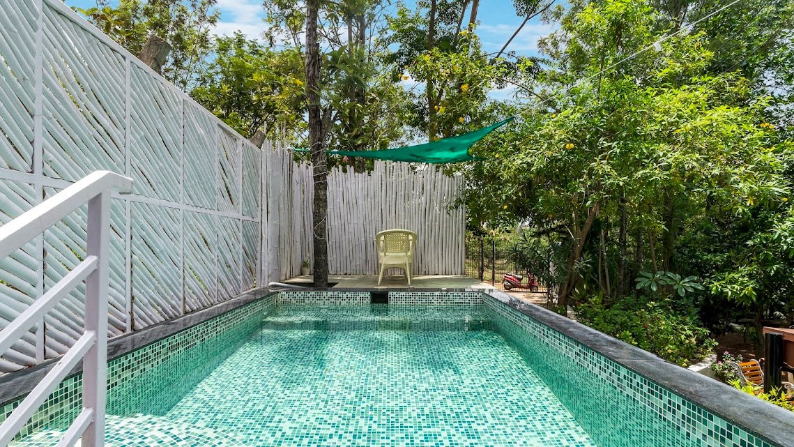 A private swimming pool is surrounded by a lush green landscape, featuring a geometric tile pattern in varying shades of green. Sunlight filters through the trees, creating dappled shadows on the water's surface. A single chair is positioned near the pool's edge.