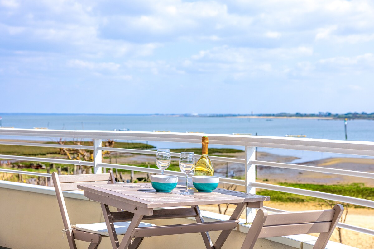 A terrace table is set with wine glasses and bowls, overlooking the sea. The horizon displays a calm waterway, with distant land visible under a cloudy sky. The arrangement invites relaxation, surrounded by the natural scenery.