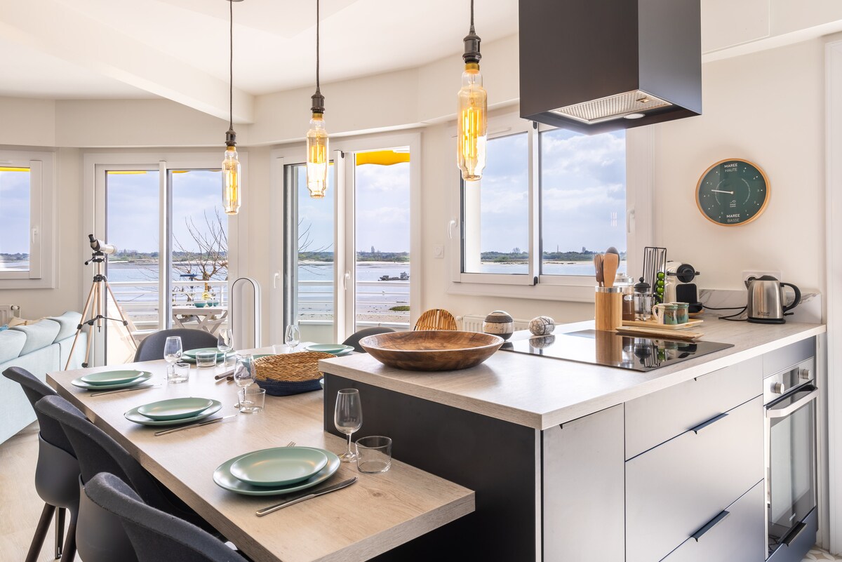 A modern kitchen featuring a spacious island with a wooden bowl at its center. The area is illuminated by pendant lights above, while large windows provide views of the surrounding landscape. A dining table set for six is visible, complemented by tableware and glassware.
