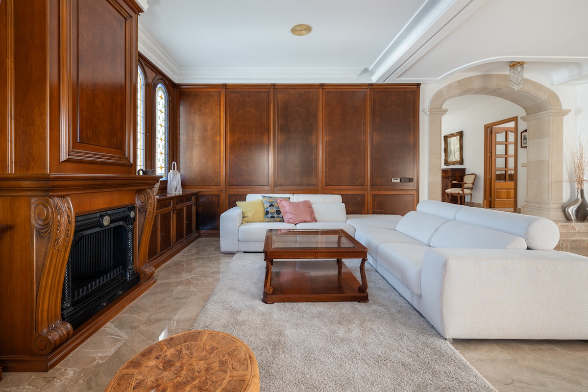 A spacious living area features a light-colored sectional sofa paired with a wooden coffee table. Natural light streams through a decorative stained glass window, illuminating the elegant wood paneling. A plush rug adds warmth to the marble floor, creating a comfortable gathering space.