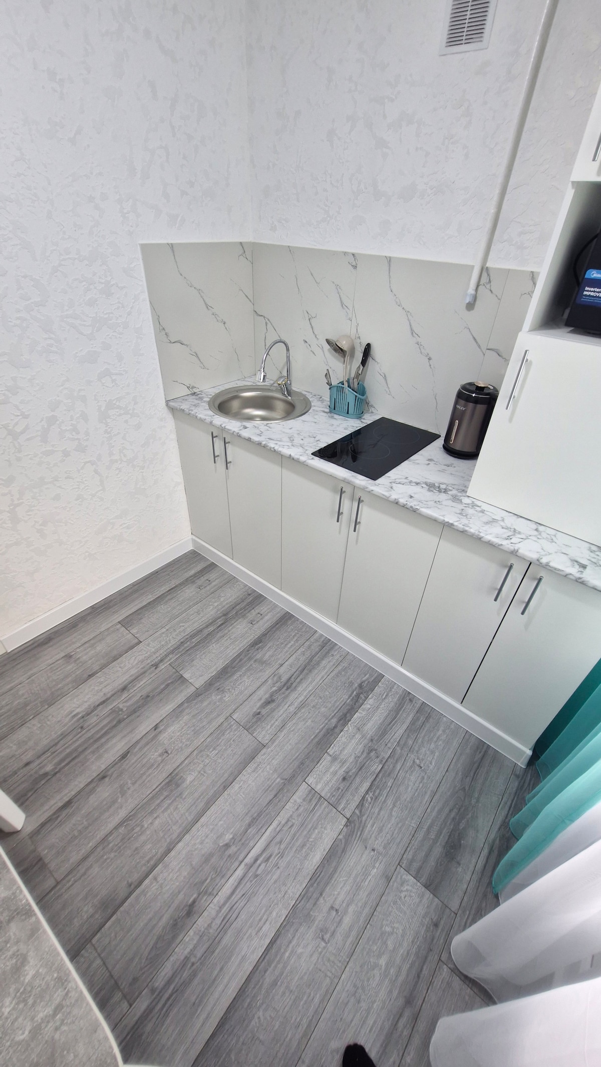 An efficiently designed kitchenette features a stainless-steel sink, an electric stovetop, and ample cabinet space. A marble countertop complements the modern aesthetic. Various kitchen utensils are visible, and a dark thermal container is placed nearby, contributing to a functional cooking area.