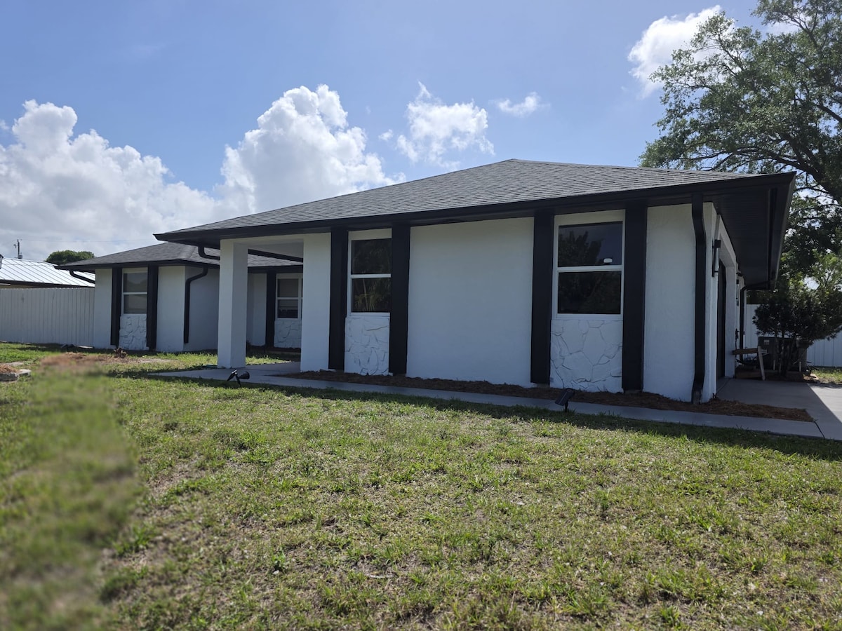 The exterior of a charming home is shown, featuring a clean white façade with dark accents. Large windows allow ample natural light, and a spacious yard is visible, bordered by lush grass. A clear blue sky and fluffy white clouds provide a serene backdrop.