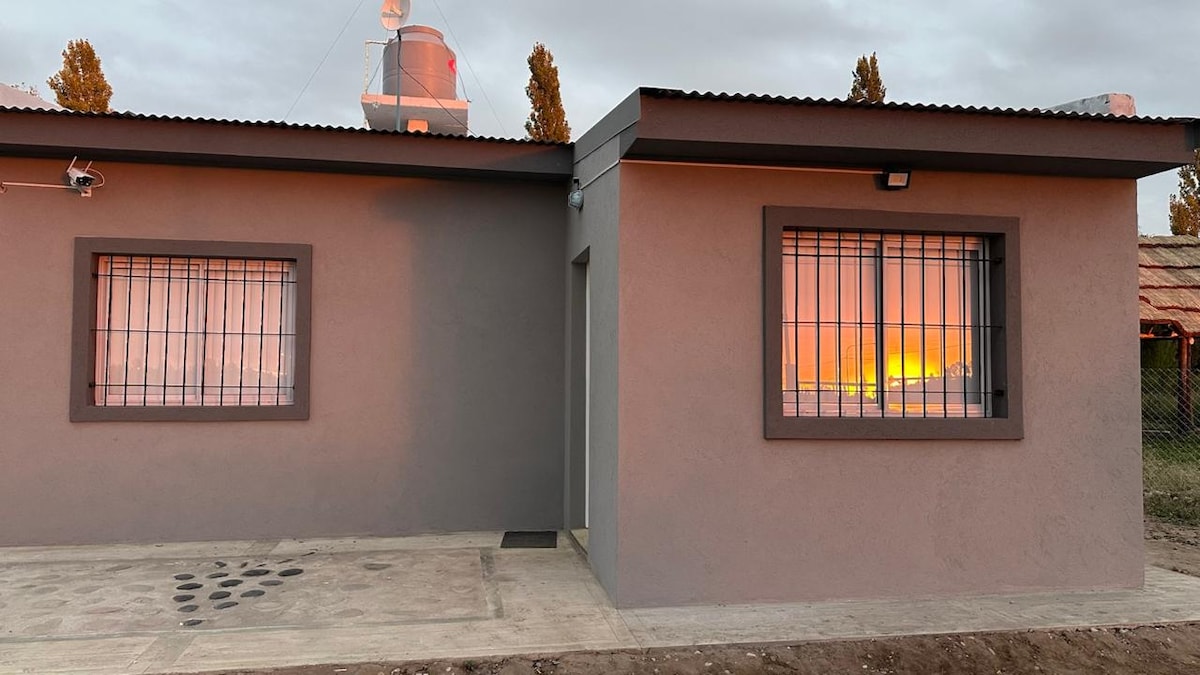 The exterior of the casita is shown, featuring a neutral-colored facade with a window framed in dark tones. The glass reflects a warm sunset glow, while a simple, clean pathway leads to the entrance. The surrounding area includes natural elements in the background.