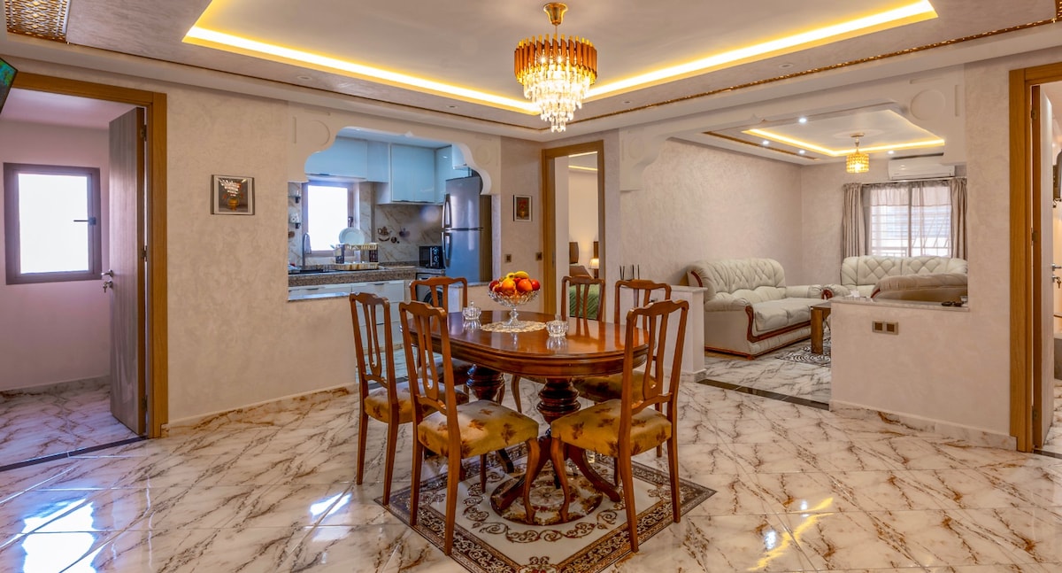 A dining area is showcased with a round wooden table surrounded by four upholstered chairs. A decorative centerpiece is present. The open layout connects to a living space featuring a light-colored sofa, with soft lighting emanating from a chandelier above, enhancing the ambiance.