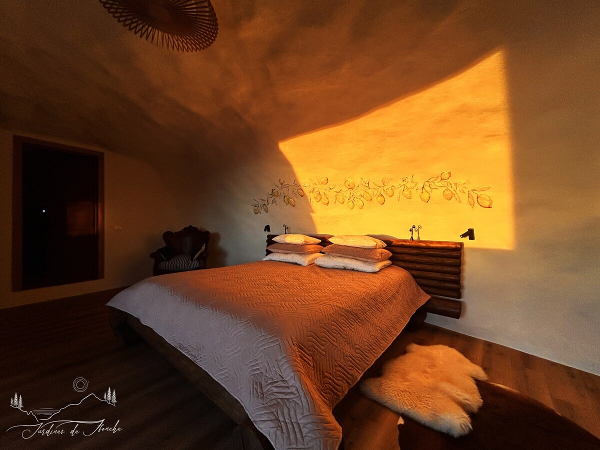 A spacious sleeping area features a wooden headboard adorned with leaf motifs, illuminated by warm evening light. The bed is dressed with neutral-toned linens and topped with fluffy pillows. A soft area rug lies beside the bed, enhancing the serene setting.