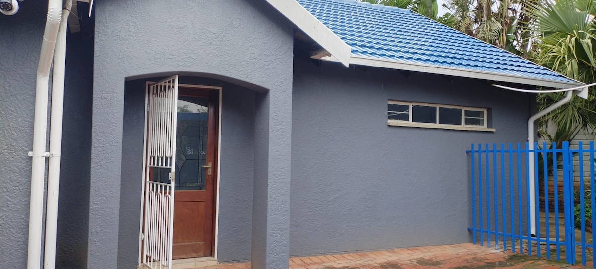 A modern exterior view of a house features a blue shingled roof and light gray walls. The entrance, framed by an arched doorway, showcases a wooden door with a security gate. A blue fence surrounds the property, with a tiled walkway leading to the entrance.
